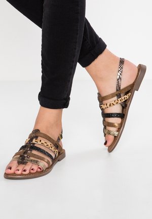 Brune og metalliske sandaler med flere remme, åben tå og teksturerede detaljer; båret med sorte bukser med opkrogede ben.