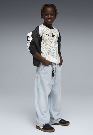 Barn med flettet hår iført en farverig stjernemønstret skjorte, sort og hvid jakke, lyseblå løse jeans og sorte sneakers med brune såler.