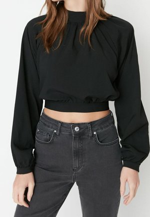 Femme portant un haut court noir à manches longues avec un col froncé et une taille cintrée, associé à un jean taille haute gris foncé.