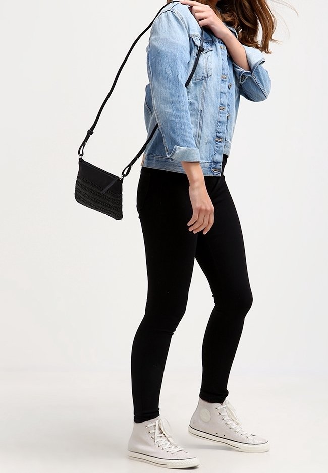 Borsa a tracolla nera con design testurizzato, appesa a una tracolla nera, indossata sopra una giacca di denim abbinata a leggings neri e sneakers bianche.