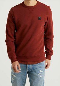 Sweat-shirt bordeaux pour homme avec poignets et encolure côtelés, présentant une petite étiquette logo noire sur la poitrine gauche. Le tissu est lisse et texturé.