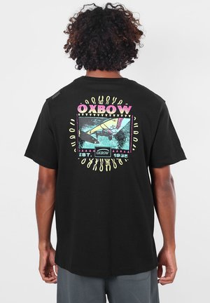 Homme aux cheveux bouclés portant un t-shirt noir avec un graphisme rectangulaire coloré de deltaplane et le texte "OXBOW EST. 1985" dans le dos.