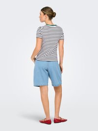 Mujer con el cabello recogido en un moño, lleva una camiseta de rayas de manga corta, pantalones cortos azul claro hasta la rodilla y zapatos planos rojos, de pie mirando hacia la izquierda.