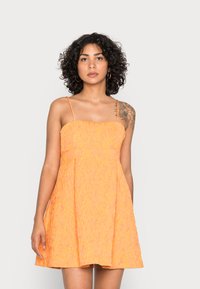 Vestido laranja, sem mangas, com um tecido texturizado, corpete justo e saia evasé. Apresenta alças finas e uma silhueta em A subtil.