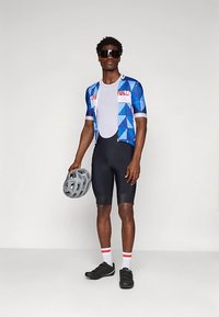 Σετ ποδηλασίας με μπλε μπλούζα γεωμετρικό σχέδιο, λευκό διχτυωτό εσώρουχο, μαύρο σορτς και μαύρα παπούτσια ποδηλασίας· κρατώντας γκρι κράνος.