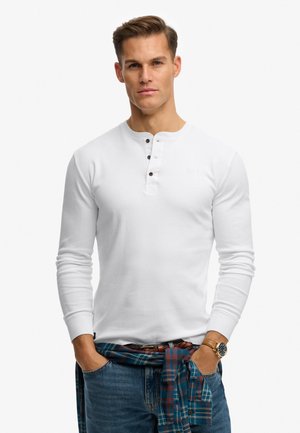 Mand iført en hvid langærmet henley skjorte, blå jeans, brun ur og ternet skjorte bundet omkring taljen.