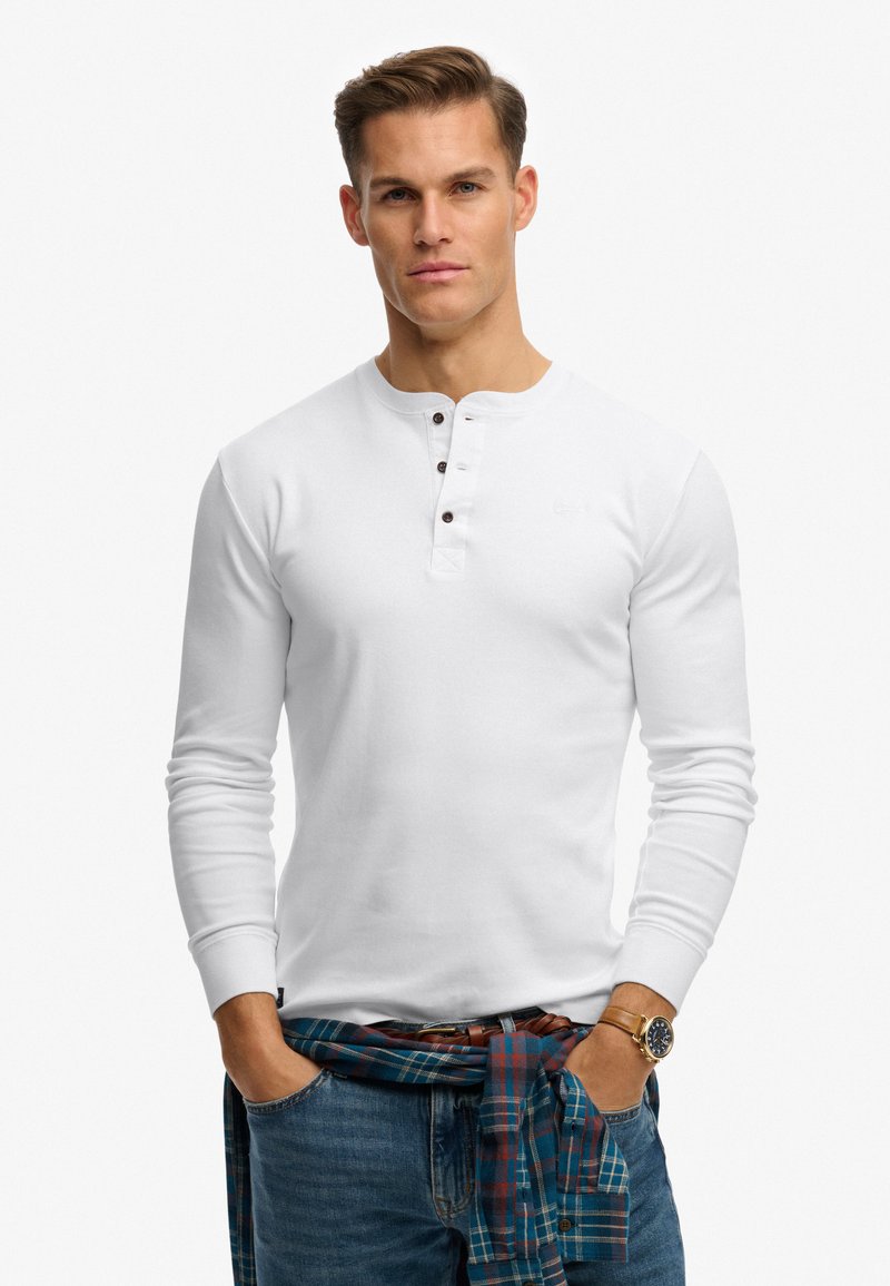 Uomo che indossa una maglia henley bianca a maniche lunghe, jeans blu, orologio marrone e camicia a quadri legata in vita.