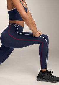 Tengerészkék leggings fehér és piros oldalsávokkal, magasított derékkal és rugalmas anyagból, fekete sportcipővel párosítva.