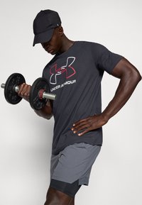 Black athletic t-shirt featuring red and white Under Armour logo, paired with gray shorts. Individual lifts a black dumbbell.