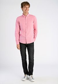 Lichtroze overhemdd met een subtiele textuur, gecombineerd met donkere jeans en witte sneakers met gekleurde accenten.