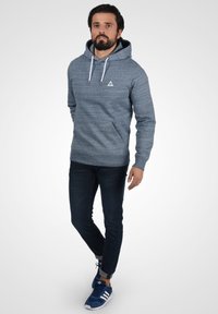 Felpa grigia con tasca frontale e logo triangolare, abbinata a jeans blu scuro e sneakers blu con strisce bianche.