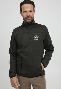 Pull-over en polaire zippé vert foncé avec une surface texturée, comportant un logo sur la poitrine, des zips rouges et des poches latérales.