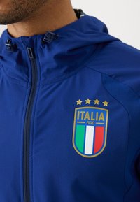 Plava lagana jakna s kapuljačom, sa zlatnim emblemom Italije i tri zvjezdice iznad njega, te teksturiranim panelima na ramenima.
