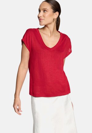 Žena s hnedými vlasmi zviazanými do copu nosí červené krátke tričko s výstrihom do V a bielu sukňu, stojaci na jednoduchom svetlom pozadí.