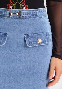 Hellblauer Jeansrock mit hoher Taille. Verfügt über zwei aufgesetzte Taschen mit goldenen Knopfakzenten und einem Metallschnallendetail am Bund.