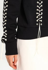 Mustat collegepaita, jossa on nauhasta solmittava muotoilu hihoissa, joissa on kermaiset läpiviennit ja nauhat, ribbireunat ja rento istuvuus.