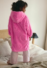 Peignoir en peluche rose avec capuche, comportant des motifs de visages souriants en relief, associé à un pantalon de pyjama rayé rose et blanc.