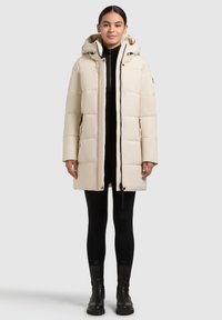 Giovane donna che indossa un cappotto trapuntato con cappuccio color crema, leggings neri e stivaletti neri, in piedi su uno sfondo neutro.