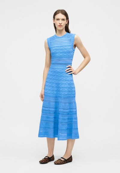 Femme debout, main sur la hanche, portant une robe midi sans manches bleue à motifs et des ballerines imprimé léopard sur fond blanc.