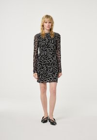 Zwarte jurk met lange doorzichtige mouwen, voorzien van een wit handgeschreven patroon. Slanke pasvorm, knielang, gemaakt van lichtgewicht stof. Zwarte schoenen.