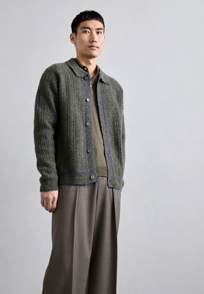 Grå ullcardigan med gröna vertikala ränder, knappstängning framtill och kragkrage. Bärs över en grön undertröja och beige byxor.
