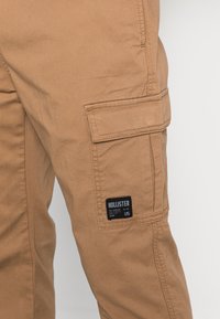 Hnědé cargo kalhoty s boční kapsou, viditelným švem a černou značkovou nášivkou s nápisem "HOLLISTER" na texturované tkanině.