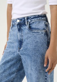 Höga, ljusblå denimjeans med en slitningseffekt och prydnader av små silvernitar över framsidan.