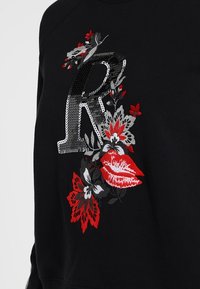 Sweatshirt noir avec un grand "R" en sequins argentés et de la broderie florale rouge, comportant une paire de lèvres rouges parmi les motifs.