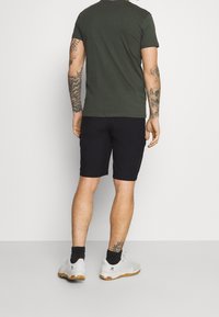 Tmavě zelené bavlněné tričko, černé kraťasy, bílé sportovní boty s texturovaným vzorem a viditelnými tetováními na paži. Jednoduchý, neformální outfit.