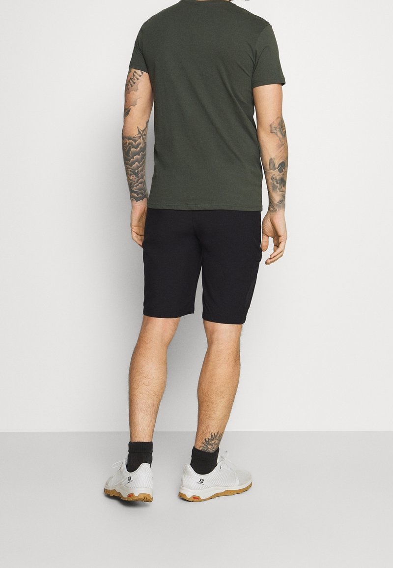 Tmavě zelené bavlněné tričko, černé kraťasy, bílé sportovní boty s texturovaným vzorem a viditelnými tetováními na paži. Jednoduchý, neformální outfit.