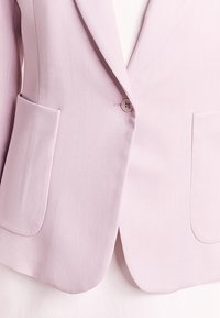 Světle fialový blazer vyrobený z hladkého materiálu; má zapínání na jeden knoflík a dvě přední kapsy s zaoblenými okraji.