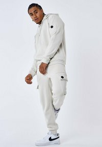 Sudadera de corte relajado en color crema con cordón, pantalones cargo a juego con bolsillos y detalles en negro, combinados con zapatillas blancas.