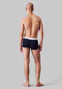 Navyblauwe boxershorts met een witte Calvin Klein tailleband, gemaakt van gladde stof, vanuit de achterkant getoond op een neutrale grijze achtergrond.