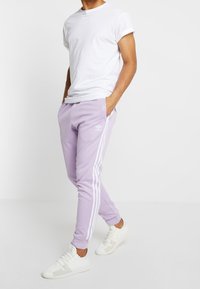 Lila sportiga joggingbyxor med vita randiga accenter, elastisk midja och sidofickor. Matchas med en enkel vit t-shirt och vita sneakers.