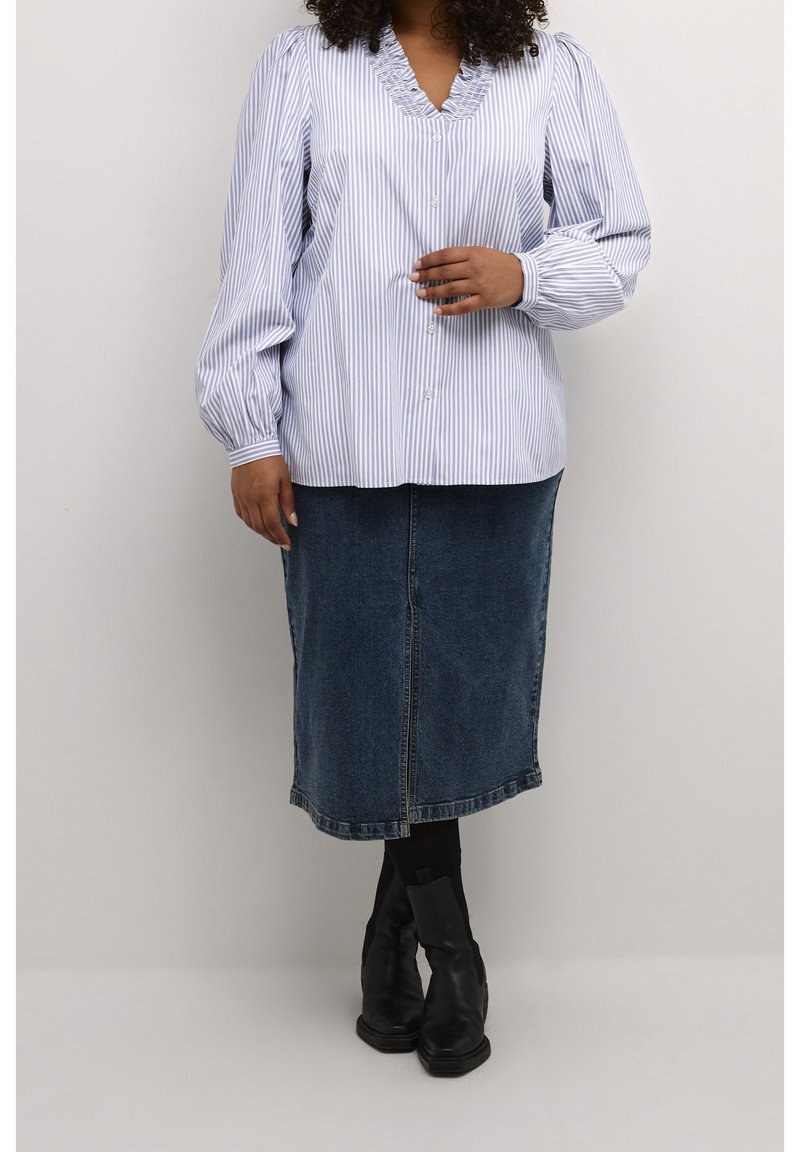 Femme portant une blouse rayée bleu et blanc à boutons, une jupe midi en jean, des collants noirs et des bottines noires, debout contre un mur uni.