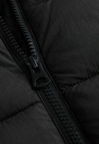 Czarny materiał kurtki puchowej zbliżenie na czarny plastikowy zamek błyskawiczny i teksturowane detale, podkreślające szwy i puchate fragmenty.