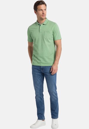 Man in een groene polo met korte mouwen en borstzak, blauwe spijkerbroek en witte sneakers, staand met ontspannen handen, kijkend naar rechts.