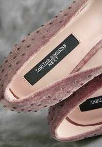 Vaaleanpunaiset samettiset tasapohjakengät, joissa on teksturoitu yläosa, jossa on pieniä pisteitä, nahkavuori ja musta tuotemerkkilappu sisäpuolella.