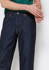 Mörkblå denimjeans med rak skärning, prydda med kopparnitar, orangeröd sömnad och femficksdesign. Tyget är slätt och hållbart.