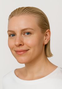 Guld diamantformad spetsörhänge på en modell med kort, slickat blont hår och en vit topp mot en ljus bakgrund.