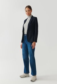 Blazer marine à deux boutons, col roulé gris et jean droit bleu associé à des chaussures de sport beiges. Tissu lisse, coupe classique.