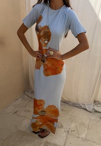 Femme portant une robe bleu clair avec de grands imprimés floraux orange, des colliers superposés, une bague en forme de soleil et des sandales noires sur un sol carrelé.