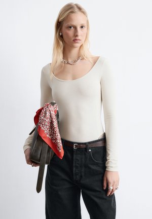 Hellbeiges Langarm-Body, kombiniert mit hochtaillierten schwarzen Jeans. Cremefarbener Schal mit rotem Blumenmuster, der an einer braunen Tasche hängt.