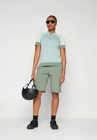 Lysegrønn sykkeltrøye med korte ermer og kvart glidelås, kombinert med lysegrønne shorts. Svart hjelm holdt i venstre hånd. Sorte sko og solbriller.