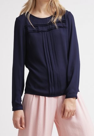 Blusa a maniche lunghe blu navy con design frontale plissettato, scollatura rotonda e tessuto morbido. Abbinata a pantaloni palazzo rosa chiaro.