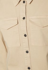 Chemise beige à boutons, fabriquée en tissu coton, avec deux poches poitrine, boutons noirs et détails de couture visibles.