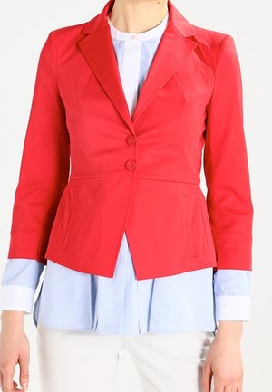 Vrouw draagt een getailleerde rode blazer met twee knopen over een lichtblauw blousehemd en witte broek, staand tegen een effen achtergrond.