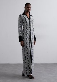 Ilga, prigludusi suknelė su zigzagų raštu pilkos, juodos ir baltos spalvos, juodomis rankovėmis ir priekyje esančia mygtukų uždarymo sistema. Išskirtinė siluetas.