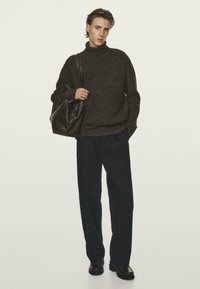 Ciemnoszary sweter z dzianiny z wysokim kołnierzem, luźny krój. Czarne spodnie o szerokich nogawkach i brązowe skórzane buty. Niesie miękką czarną torbę. Neutralne tło.