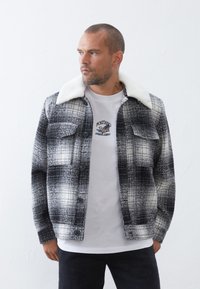 Giacca a quadri in bianco e nero, con colletto in pelliccia sintetica, chiusura a bottoni e due tasche sul petto, indossata sopra una t-shirt bianca con grafica.